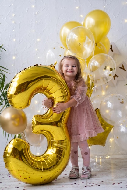 Happy little blond girl in pink dress holding big gold foil balloon number and digit three