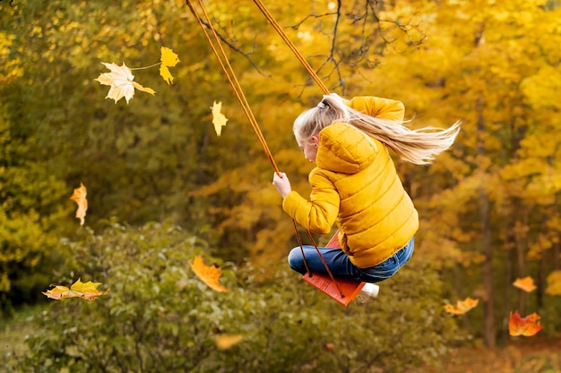 Happy little blonde caucasian girl smiling and riding a rope swing in autumn in the park