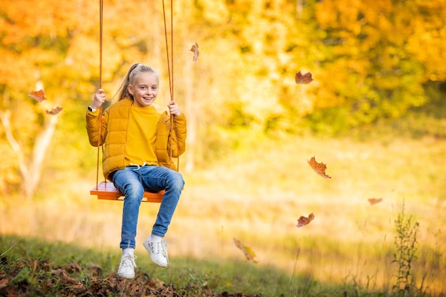 Happy little blonde caucasian girl smiling and riding a rope swing in autumn in the park