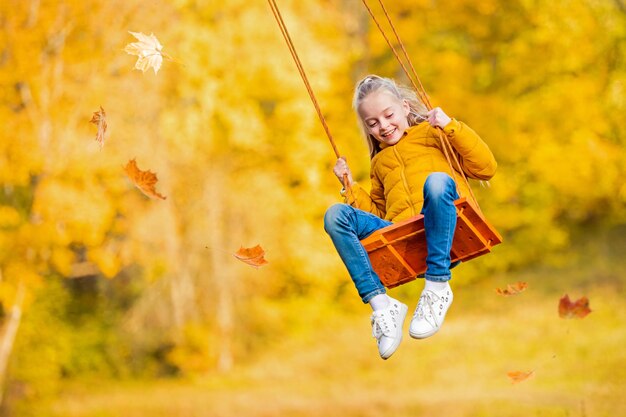 happy little blonde caucasian girl smiling and riding a rope swing in autumn in the park