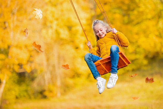 Happy little blonde caucasian girl smiling and riding a rope swing in autumn in the park