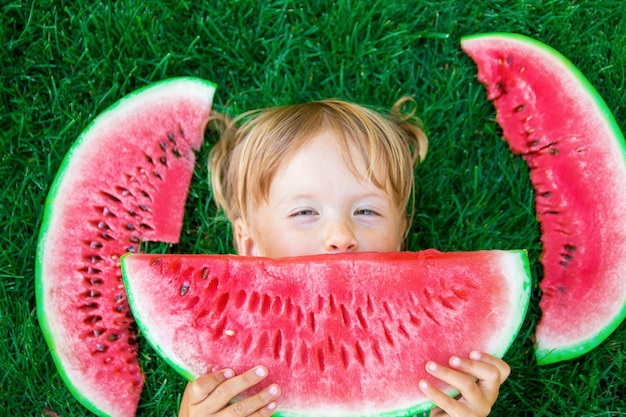 Happy little blonde girl lying on the grass with big slice watermelon in summer time.