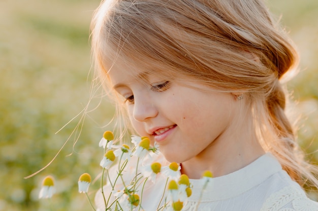 Happy little blonde girl sniffs a bunch of daisies in summer at sunset