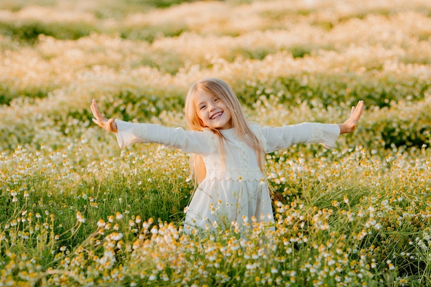 Happy little blonde girl in white cotton dress raised her hands up in a field of daisies