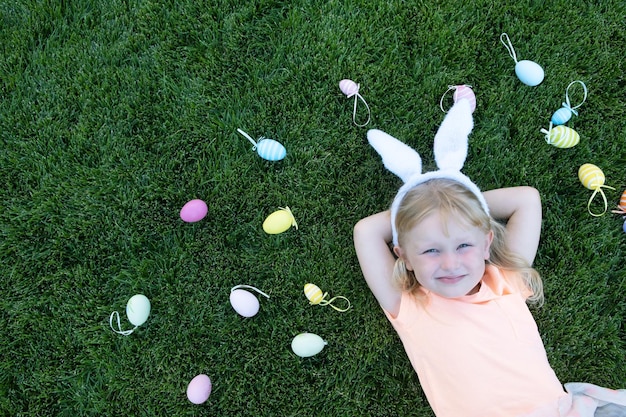 Foto felice bambina bionda con orecchie da coniglio sdraiato su un'erba tra colorate uova di pasqua vacanze di pasqua