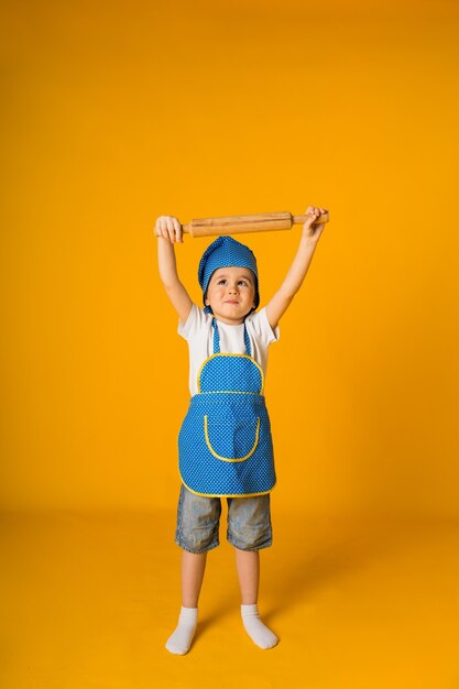 Happy little boy in a chef's costume holds a wooden rolling pin on a yellow surface with space for text