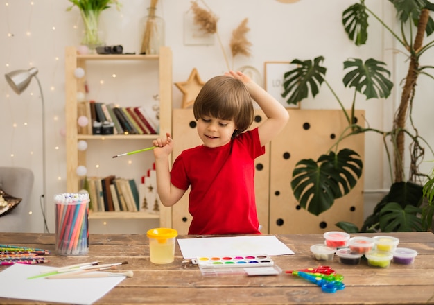 Happy little boy draws with a brush and paints on white paper on a wooden desk with stationery