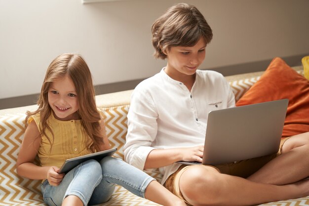 Happy little boy and girl brother and sister sitting on sofa at home using laptop and digital