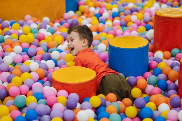 행복한 어린 소년은 형형형색색의 공이 있는 마른 수영장에 뛰어드는 것을 즐긴다 그 꼬마 소년은 형형형색색의 공이 있는 게임방에서 놀고 즐거운 시간을 보내고 있다