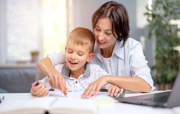 幸せな男の子と彼の思いやりのある母親は教育のために家で時間を過ごす魅力的な女性と彼女の息子は机に一緒に座ってコピーブックに書いています