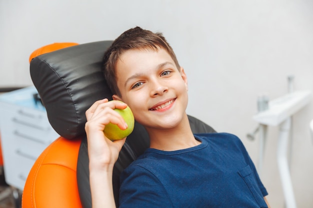 Happy little boy holding an apple while sitting in the dentist's chair and looking at the camera