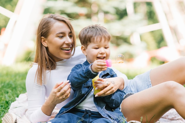 幸せな小さな男の子は、彼の幸せな若い母親と一緒にシャボン玉を吹いて遊んでいます 週末の休憩 思いやりのある子育てと育児