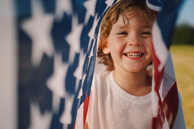 幸せな少年愛国者がアメリカ国旗を持って野原を走っている。アメリカは 7 月 4 日を祝う