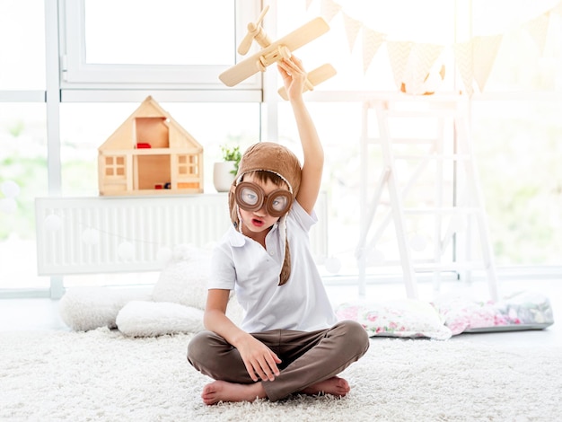 Happy little boy in pilot hat playing with wooden plane in children's room