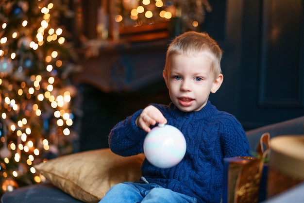 青いニットの居心地の良いリビングルームでクリスマスツリーの装飾が施されたソファに座っている幸せな少年...