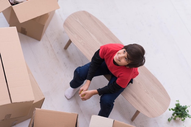 счастливый маленький мальчик сидит на столе с картонными коробками вокруг него в новом современном доме, вид сверху