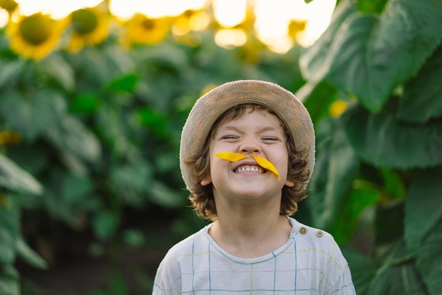 Happy little boy walking in field of sunflowers and making a mustache from sunflower petals