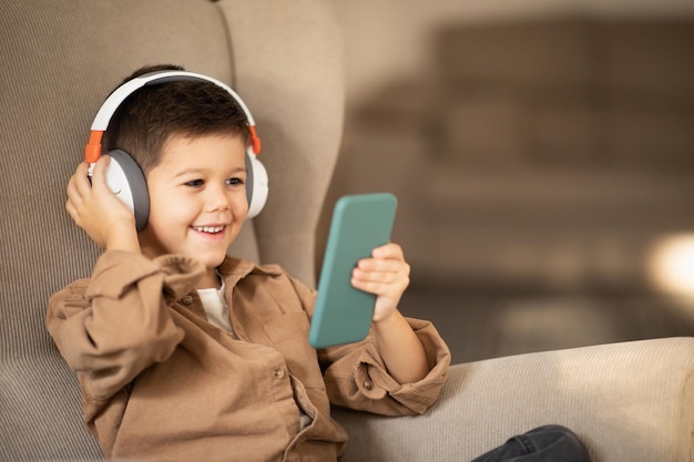 ワイヤレスヘッドフォンで幸せな小さな男の子がスマートフォンでビデオを見る電話で肘掛け椅子に座っている