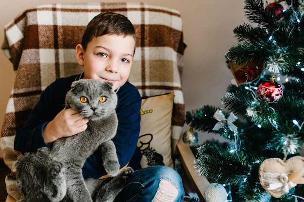 猫と幸せな小さな男の子-自宅のクリスマスツリーの近くに座っているクリスマスプレゼント。メリークリスマスとハッピーホリデー！屋内でかわいい子。