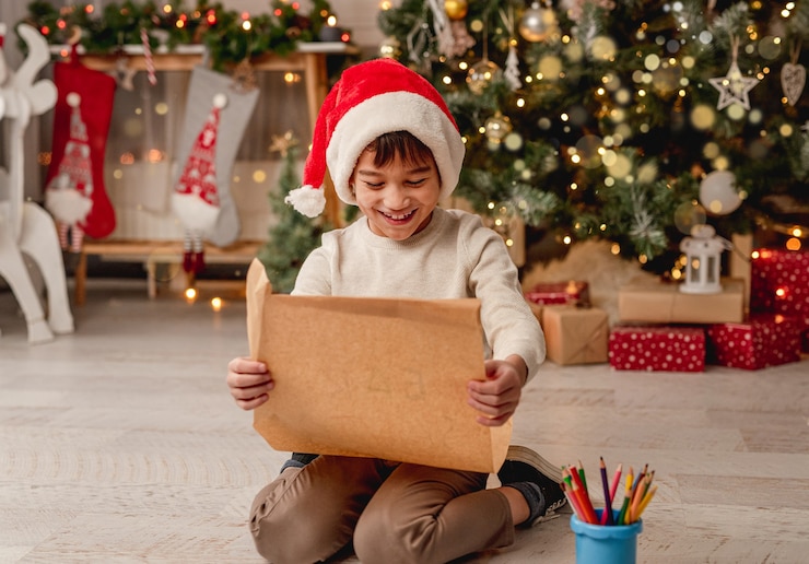 Photo happy little boy with letter to santa sitting on floor in festive room with christmas tree