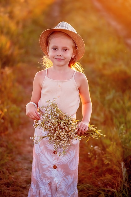 Happy little caucasian girl 5 years old with a bouquet of flowers of chamomiles posing in the meadow looking at the camera, a portrait at sunset