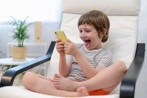 Happy little child boy playing online game watching video on smartphone sitting on chair