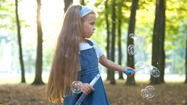 緑の公園の外でシャボン玉を吹く幸せな小さな子の女の子。子供のための屋外の夏の活動の概念。