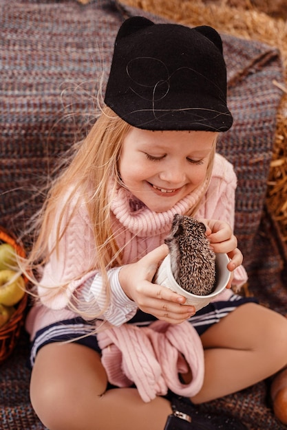 Happy little child girl with cute hedgehog Portrait of kid with pet in autumn nature with pumpkins and straw holding a cup of hot drink