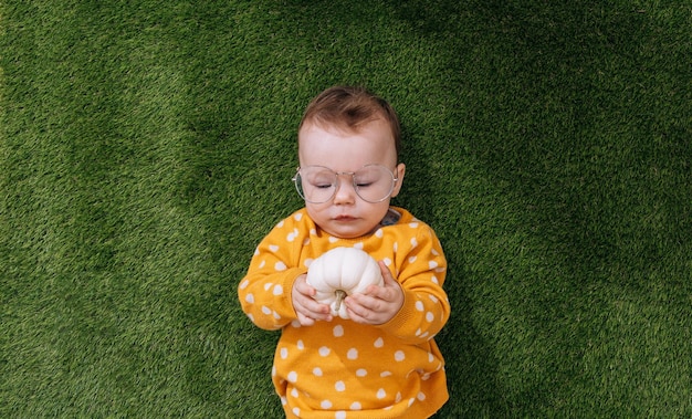 A happy little child lies on the green grass in a yellow sweater and glasses holds a pumpkin in his hands