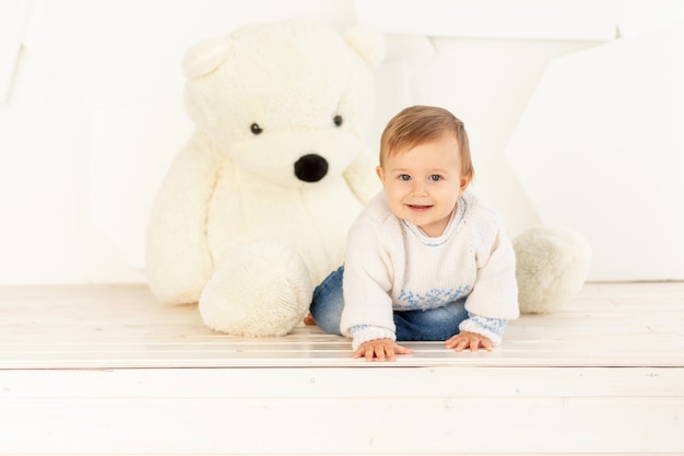 Un piccolo bambino felice di sei mesi in una giacca calda lavorata a maglia e jeans blu striscia a casa vicino a un grande orsacchiotto