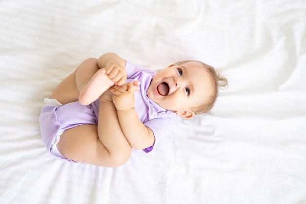 Happy little cute girl child in a bodysuit lies on her back on a bed on white linens, smiles, plays with her legs. The concept of baby products.