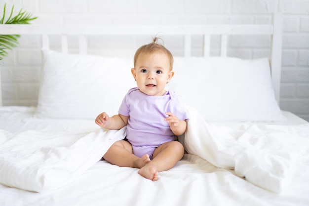 Happy little cute girl child in bodysuit sits on bed on white\
bedding smiling the concept of baby products