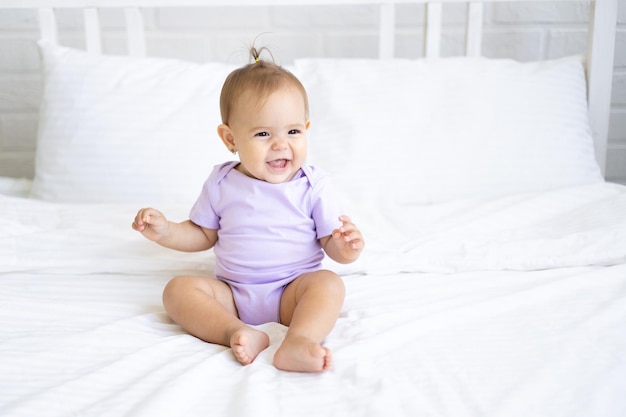 Happy little cute girl child in bodysuit sits on bed on white bedding, smiling. The concept of baby products.