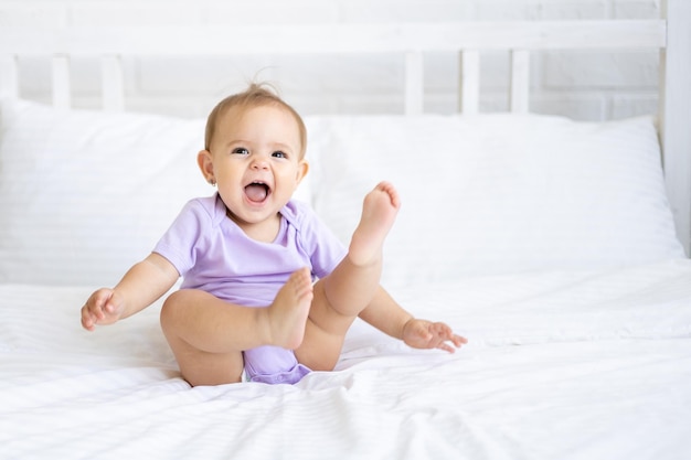 Happy little cute girl child in bodysuit sits on bed on white bedding, smiling. The concept of baby products.