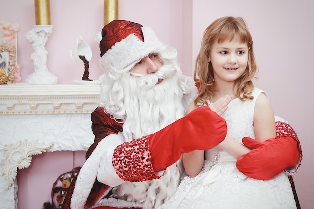 Happy little cute girl sitting on the lap of aged Santa Claus and hugs at home near christmas tree