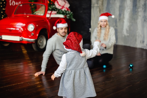 写真 サンタの帽子をかぶった幸せな小さな娘は、クリスマスイブにクリスマスツリーの近くのお母さんとお父さんに行きます。家族、子供時代、休日、人々の概念。