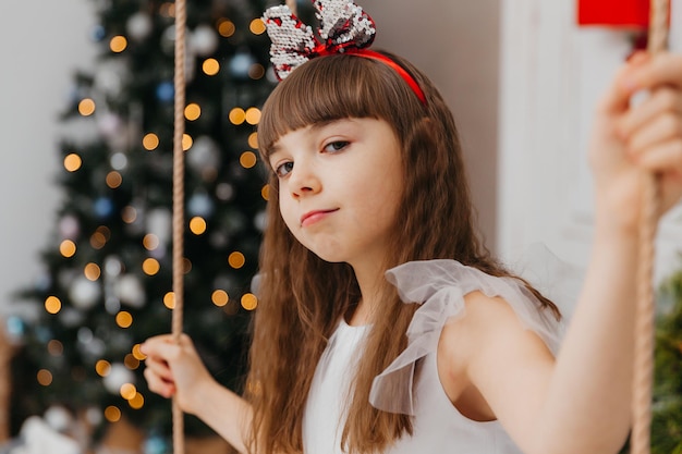 Happy little girl in a beautiful white dress sits on a swing near a Christmas tree in the studio. New Year.