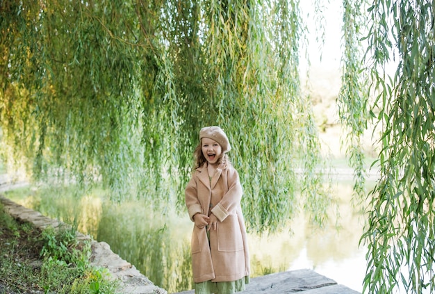 a happy little girl in a beige coat and beret is standing near a willow tree