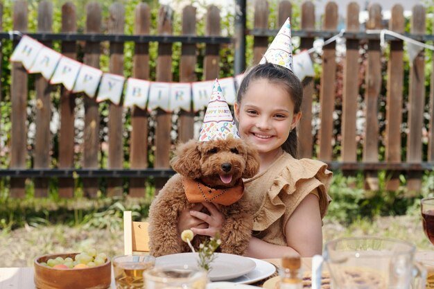 Happy little girl in birthday cap holding cute pet dog while sitting by table