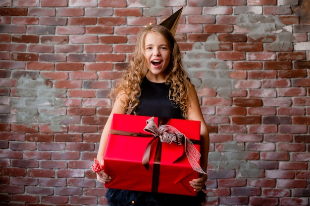 Happy little girl in a birthday cap holds a large red gift box.