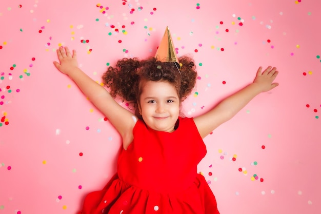 Happy little girl in a birthday cap lies among the multicolored confetti on a pink background a view from above