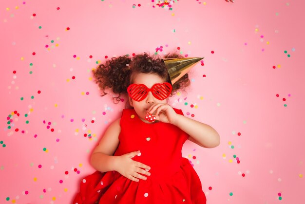 Happy little girl in a birthday cap lies among the multicolored confetti on a pink background a view from above