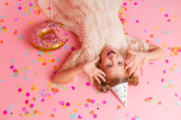 Happy little girl in a birthday cap lies among the multicolored confetti on a pink background