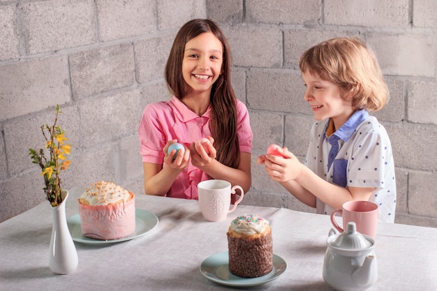 Happy little girl and boy playing traditional Easter game - egg tapping with colored eggs at home