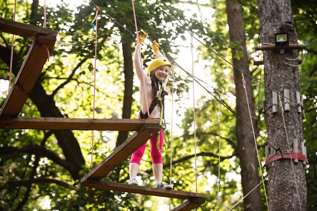 나무를 오르는 행복한 소녀 Go Ape Adventure Hike 및 어린이 개념 나무 위의 집이나 밧줄 공원에서 안전 장비를 등반하는 귀여운 소녀가 밧줄을 오르는