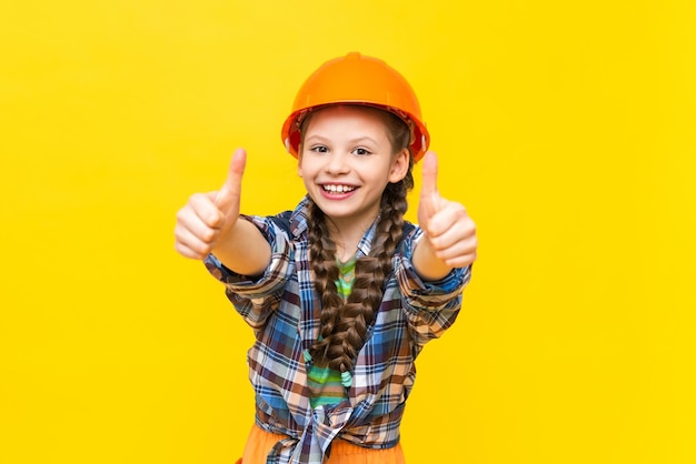 A happy little girl in a construction helmet and shirt on a yellow isolated background The child is preparing for repairs in the nursery and gives a thumbs up Yellow isolated background