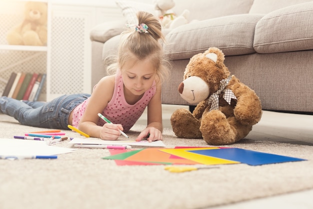 Happy little girl drawing. Cute kid lying at home on floor among colored paper and pencils. DIY, creative art hobby, early development and inspiration concept