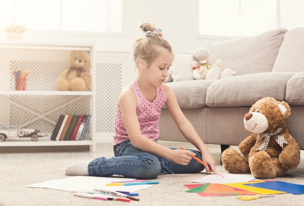 Happy little girl drawing. Cute kid sitting at home on floor among colored paper and pencils. DIY, creative art hobby, early development and inspiration concept