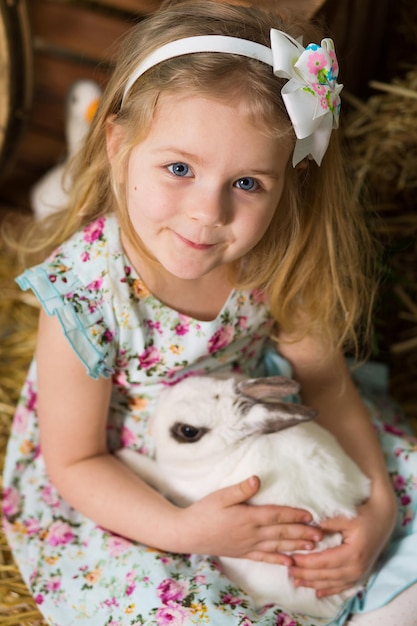 A happy little girl in a dress sits in a nest and holds a cute fluffy white Easter bunny