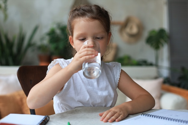 Happy little girl drinks crystal still mineral water in glass, small child recommend daily dose of clean aqua